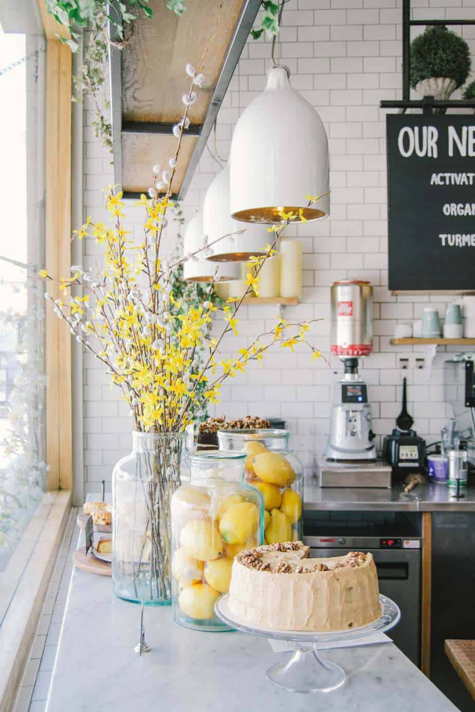 The white kitchen counter with a clear glass vase filled with yellow flowers and a cake on a stand with a piece taken out of it.