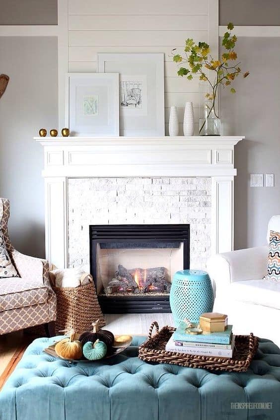 A white fireplace mantel with a baby blue ottoman in front of the fireplace.   There is a clear glass vase with a branch and green leaves on it.