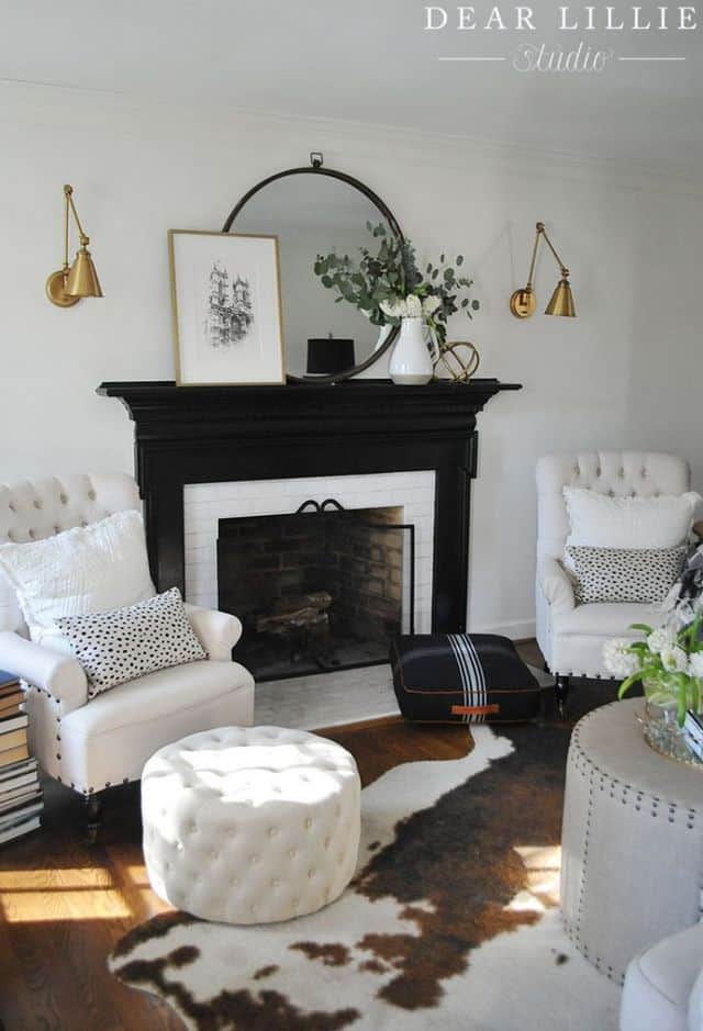 A black fireplace with gold sconces flanking it.  There are two white armchairs on either side with throw pillows on them.   A cow hide rug is in front with a white ottoman.
