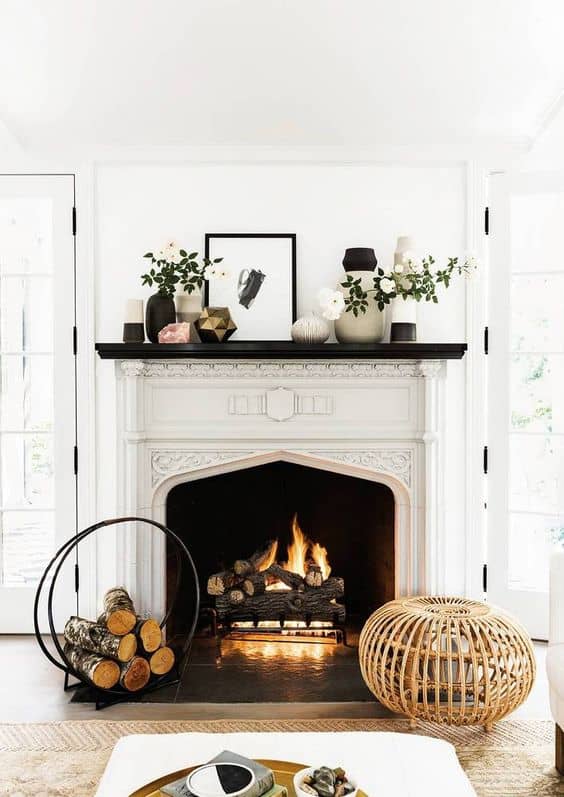 A lit fire is in the fireplace, there are vases filled with flowers and greenery and a black and white picture sits on the mantel.