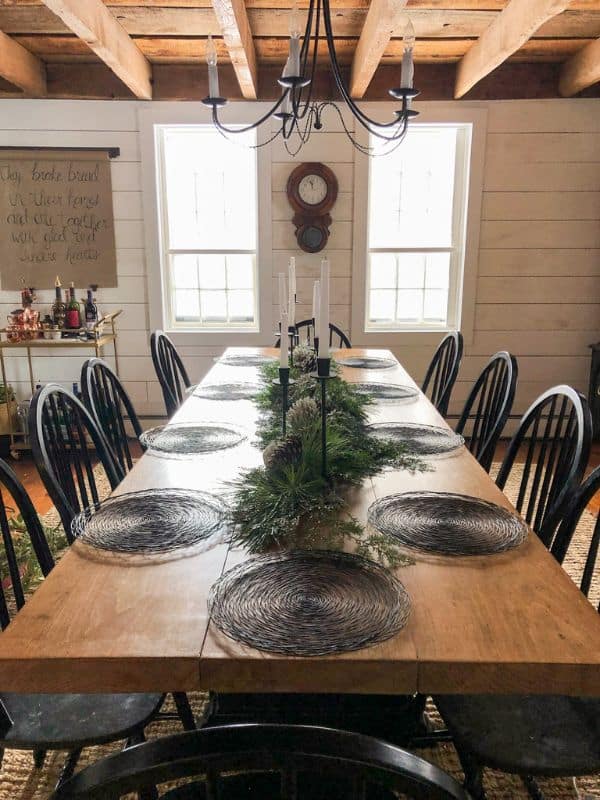 Dining table with black chargers as the table setting, fresh garland down the center of the table, and candle sticks and pinecones onto of the garland.