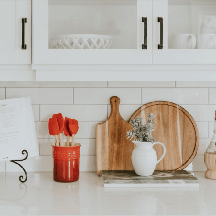 A mostly white kitchen with a red ceramic container with kitchen tools in it, plus a variety of wooden cutting boards and a white pitcher filled with greenery.