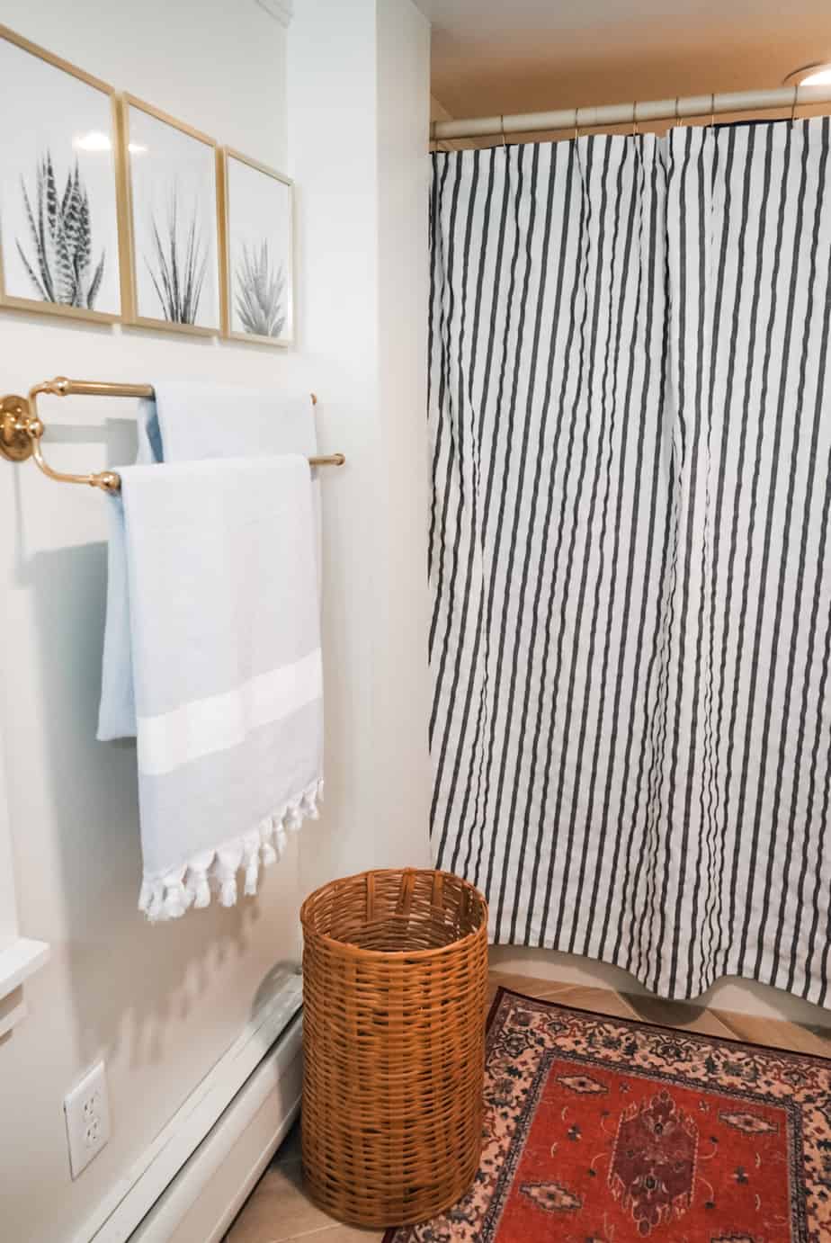 Blue and white shower curtain and towels, with three pictures on the wall and a wicker clothing hamper underneath the towel rack.