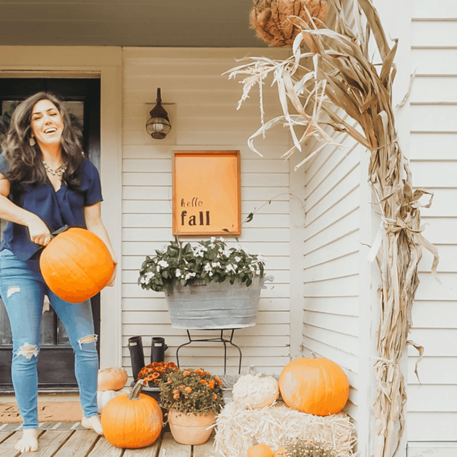 a woman holding a a pumpkin on the front porch with pumpkin, mums, and corn stalks all around her