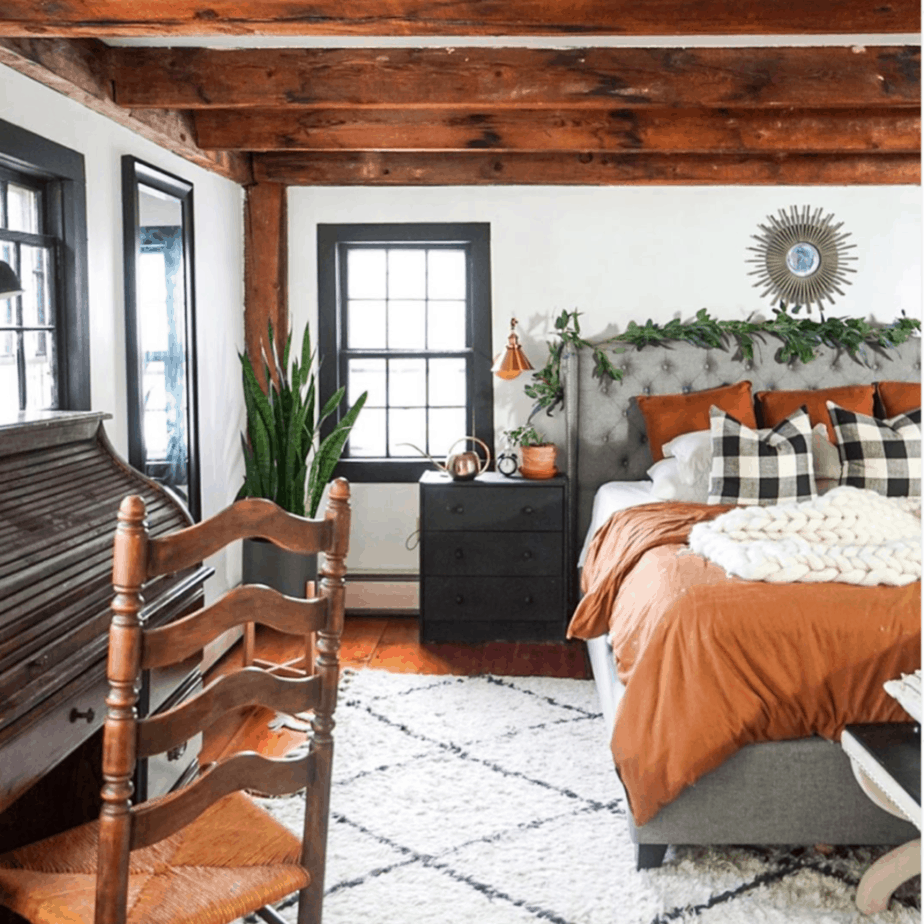farmhouse bedroom with exposed wooden beams and rust colored bedding. wooden secretary desk and wooden chair. buffalo plaid pillows and white knit blanket on bed.